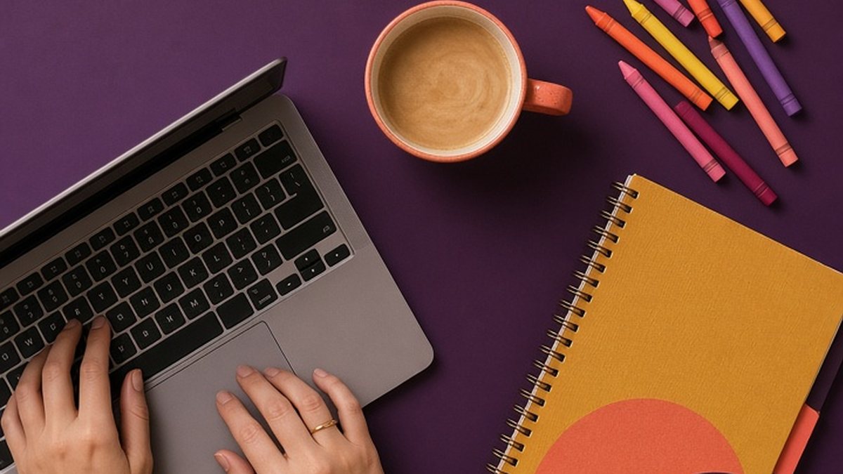 Woman's hands typing on a laptop on a plum desk next to coffee, crayons and a mustard notebook