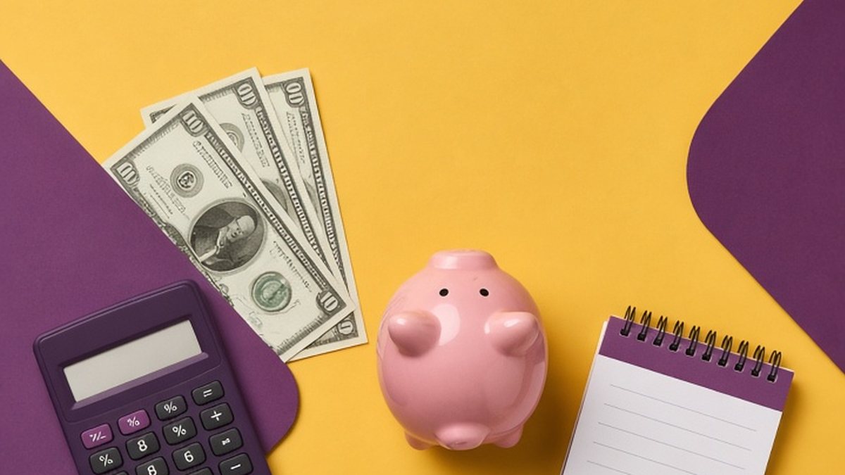 Calculator, dollar bills, pink piggy bank and blank notepad on a mustard and plum background